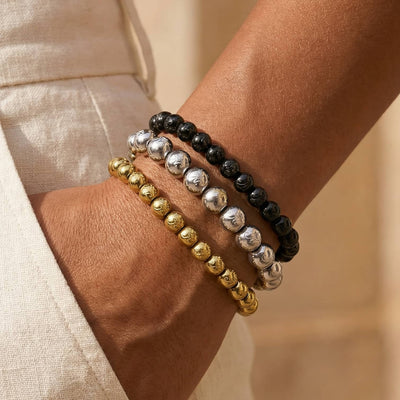 NOGU Essentials + Vault Unlock stack of gold, silver, and black beaded stretch bracelets worn on the wrist.