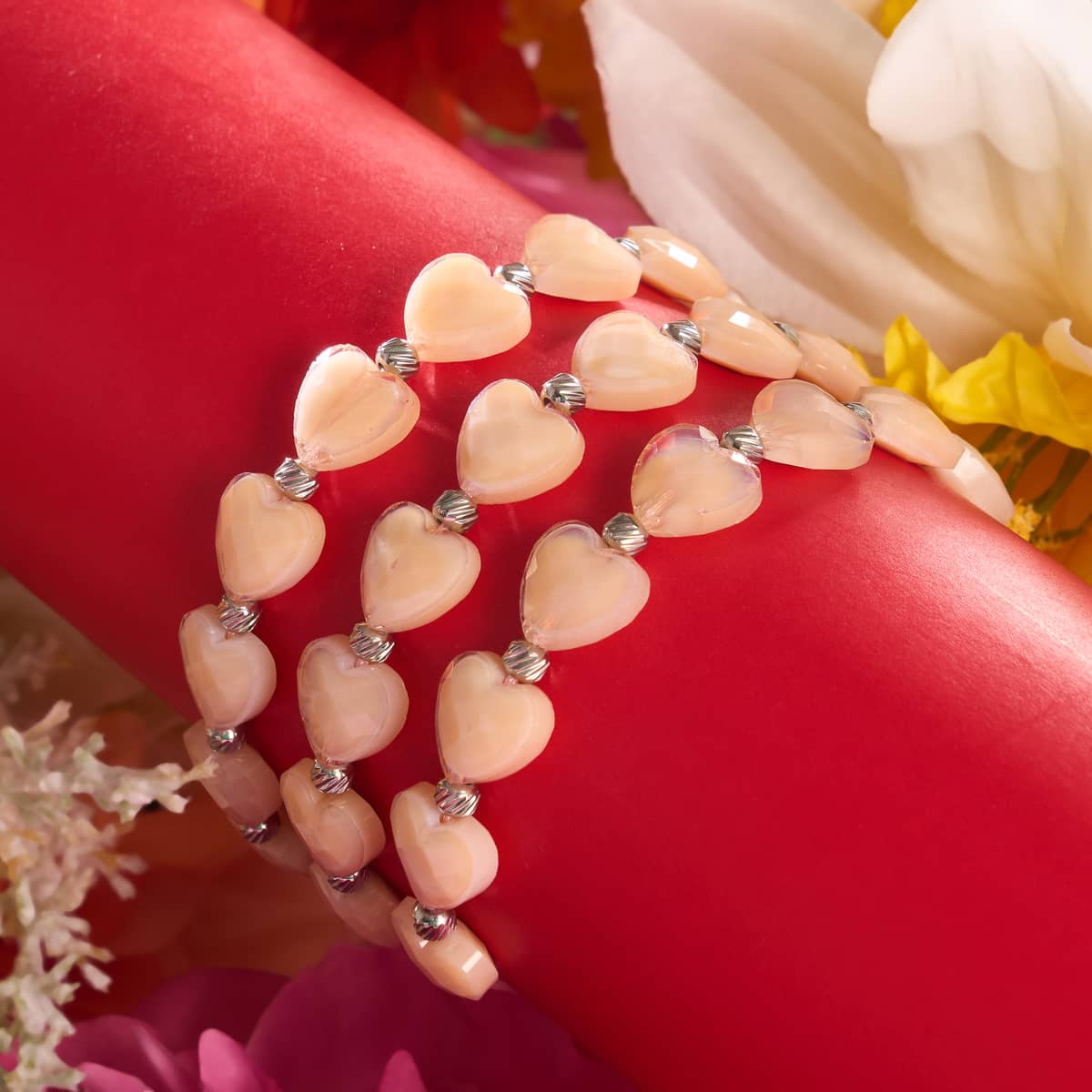 Rosé Heartstring crystal macrame bracelet with champagne heart beads and silver accents displayed on red surface surrounded by flowers by NOGU.