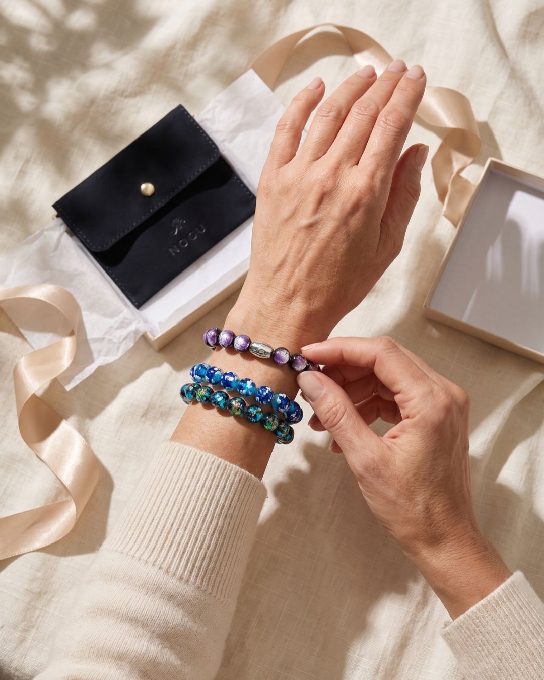 Woman wearing stacked blue and purple beaded stretch bracelets with a silver-toned accent bead beside NOGU gift packaging.